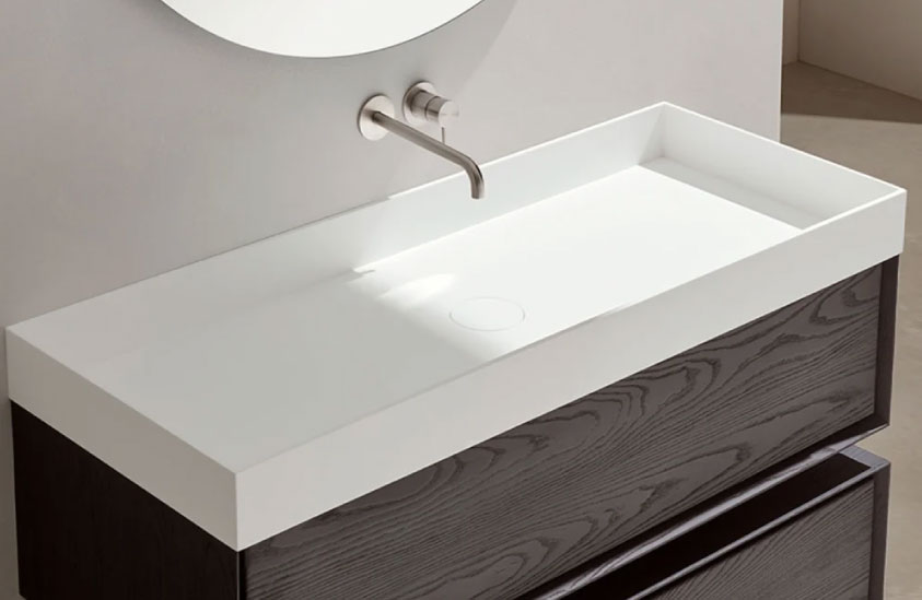 Mueble de baño color madera oscuro y lavabo color blanco. Grifería en plata empotrada en la pared y espejo circular.